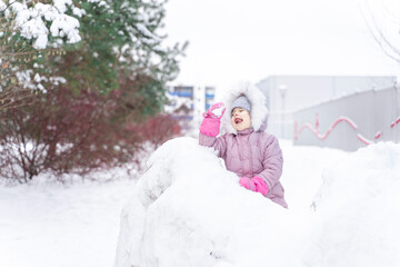 Fototapeta na wymiar little girl in a warm coat and a hood plays snowballs in a snow fortress. active winter weekend, seasonal outdoor activities, happy family lifestyle