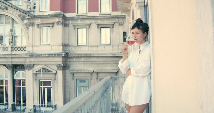 Cute woman drinking a glass of rose wine in a balcony wearing only a shirt. European lifestyle.