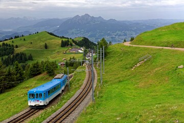 Naklejka na ściany i meble A sightseeing train traveling on the cogwheel railway through grassy alpine meadows on Mt. Rigi, with rugged Pilatus among majestic Alpine mountains in background on a cloudy summer day in Switzerland
