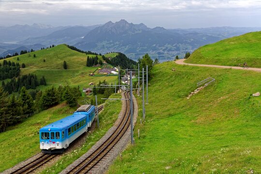 A sightseeing train traveling on the cogwheel railway through grassy alpine meadows on Mt. Rigi, with rugged Pilatus among majestic Alpine mountains in background on a cloudy summer day in Switzerland
