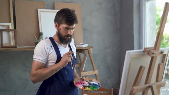 A young bearded Caucasian male painter dressed in a blue apron stands near an easel with a canvas holding a palette with paints in his hands and thinks about what to draw.