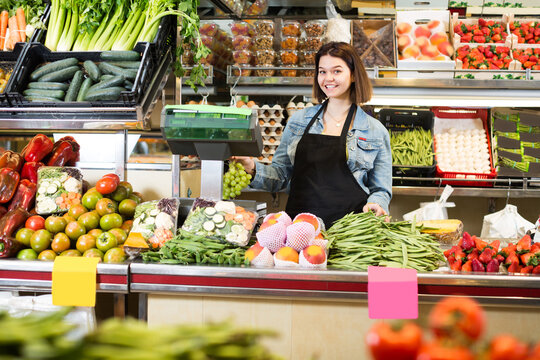 Smiling girl seller is weighing fruit and vegetables in grocery shop.