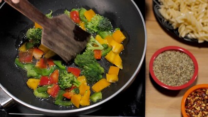 Capsicum broccoli red and yellow bell peppers being stir-fried in a pan. Closeup shot of oregano and chili flakes kept with a plate of pasta on a wooden table - popular Italian recipe - Powered by Adobe