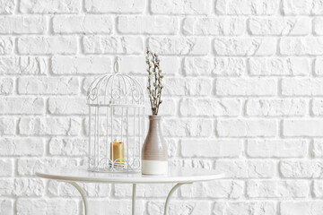 Vase with willow branches and burning candle on table near brick wall