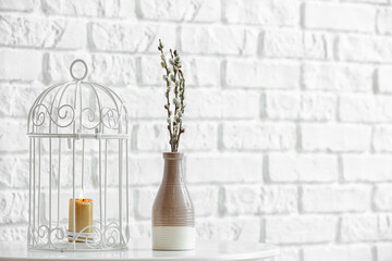 Vase with willow branches and burning candle on table near brick wall