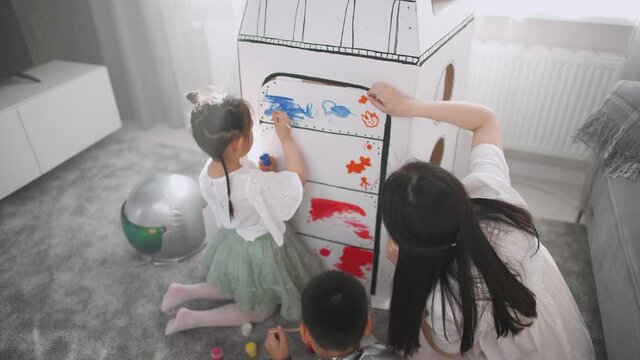 An Asian female with kids play in the living room at home, a boy in an astronaut costume sitting on the floor with her mother and sister, children together with their mother paint on a cardboard model