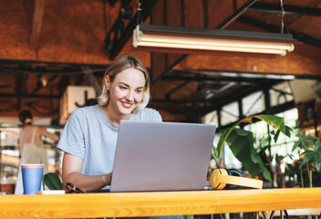 Young smiling blonde woman freelancer in yellow headphones working on laptop on table at cafe