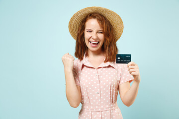 Sutisfied smiling young redhead curly woman 20s hold in hand credit bank card do winner gesture wears casual pink dress straw hat look camera isolated on pastel blue color background studio portrait