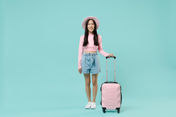 Full size body traveler tourist asian woman wear pink casual clothes hat hold suitcase bag isolated on pastel pink color background studio. Passenger travel abroad getaway. Air flight journey concept.
