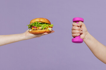 Close up young female arm hand holding violet dumbbells and junk fast food hamburger cheeseburger burger isolated on purple color background home gym Unhealthy nutrition diet sport motivation concept.