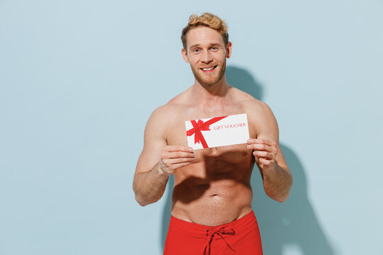 Handsome young sexy man in red shorts swimsuit relax near hotel pool hold gift certificate coupon voucher card for store isolated on pastel blue background. Summer vacation sea rest sun tan concept.