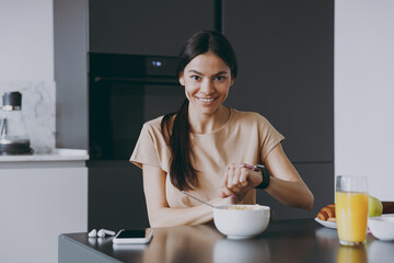 Young smiling happy punctual housewife woman 20s in beige t-shirt eat breakfast in morning look camera use smart watch check time cooking food in light kitchen at home Healthy diet lifestyle concept