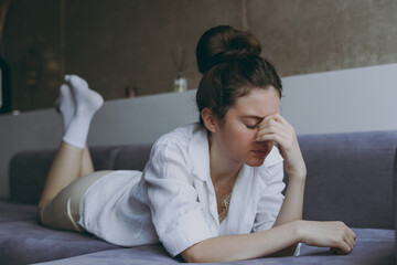 Full length young sad thoughtful depressed puzzled pensive woman 20s in white clothes lying on soft grey sofa indoors apartment put hand on nose Rest on weekends leisure quarantine stay home concept.