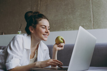 Young side view smiling copywriter freelancer woman in white clothes lying on grey sofa indoor...