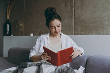 Young calm happy woman 20s in white clothes plaid sitting on soft grey sofa indoors apartment hold glass drink red wine read novel book textbook Rest on weekend leisure quarantine stay home concept.