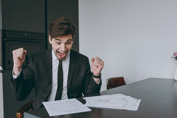 Young overjoyed employee business man corporate lawyer 20s wearing formal black suit shirt tie read paper documents do winner gesture clench fist sit at table in kitchen. People lifestyle concept