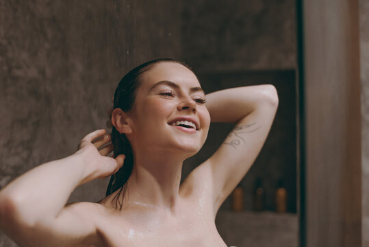 Smiling half naked topless young wet haired woman 20s take hot evening shower stand under water in bathroom do morning routine in brown background. Skin care healthcare cosmetic procedures concept