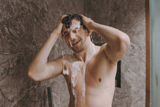 Young handsome half naked topless relaxed brunette caucasian man 20s taking hot shower washing hair with shampoo indoors in bathroom at home. People healthcare daily morning routine lifestyle concept