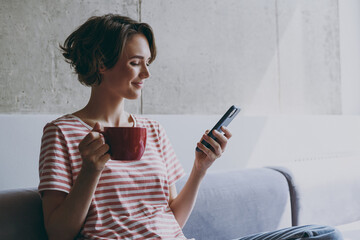 Young caucasian smiling happy woman 20s in casual clothes sitting on grey sofa using mobile cell...