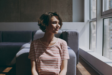 Young minded dreamful calm happy caucasian woman 20s in casual clothes sitting near grey sofa look...