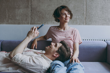 Young playful couple two friends woman man 20s in casual clothes sitting on sofa boyfriend lying on girlfriend use mobile phone look aside rest indoors at home flat together. People lifestyle concept.
