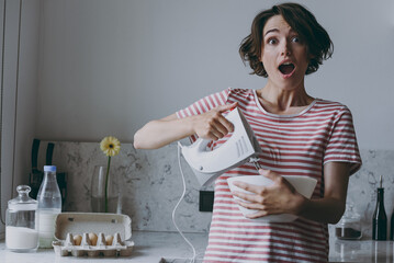 Young excited surprise brunette housewife woman in casual clothes striped t-shirt using mixer whips...