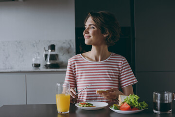 Young smiling housewife woman in casual clothes striped t-shirt eat vegeterian breakfast lunch vegetable salad look aside cooking food in light kitchen at home alone Healthy diet lifestyle concept