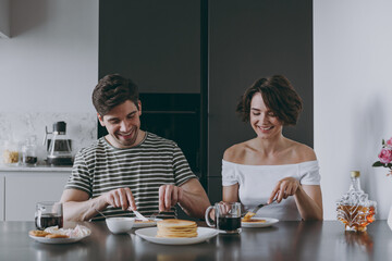 Young cheerful happy couple two woman man in casual t-shirt clothes sit by table eat pancake with maple syrup have breakfast cook food in light kitchen at home together Healthy diet lifestyle concept