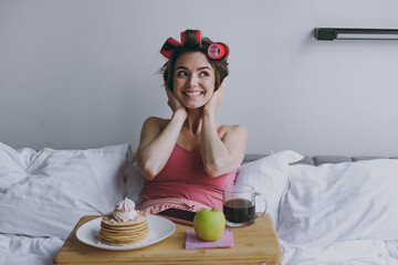 Surprised fun dreamful amazed happy young woman wear pajamas hair curlers lying in bed wrap covered...