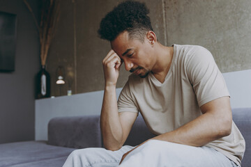 Side view pensive thoughtful confused young african american man in beige t-shirt sit on grey sofa indoors apartment procrastinate prop up forehead rest on weekends stay at home Tattoo translate fun.