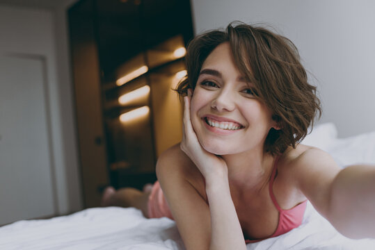 Close up young woman 20s wearing pajamas pink tank top shirt lying in bed do selfie shot on mobile cell phone rest relax indoors at home show thumb up gesture Good mood night morning bedtime concept