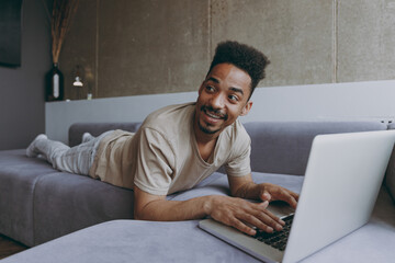 Young excited happy fun joyful african american man wearing beige t-shirt sweatpants lay down on grey sofa indoors apartment use laptop pc computer look aside resting on quarantine weekends stay home.