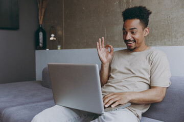 Young cool african american man in casual beige t-shirt sweatpants sitting on grey sofa indoors...
