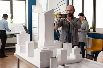Architect profession man looking at maquette layout using smartphone to design building model plan...