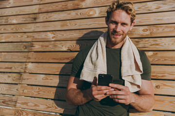Young strong sporty athletic fit sportsman man in sports clothes towel warm up training hold mobile cell phone typing message leaning on wooden wall sea beach outdoor seaside in summer day morning