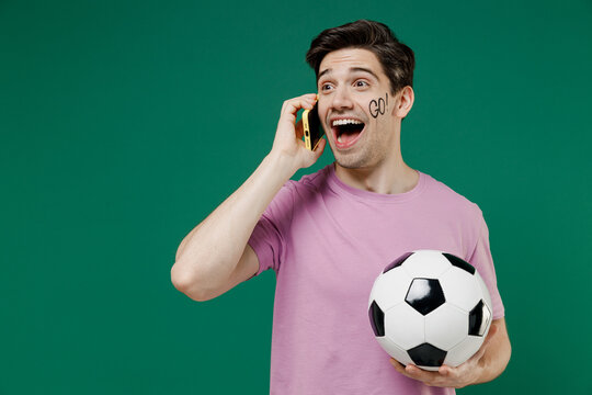 Young fun man fan wear basic pink t-shirt cheer up support football sport team hold in hand mobile cell phone soccer ball watch tv live stream scream isolated on dark green color background studio
