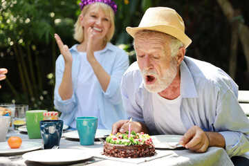 Happy multiethnic family giving surprise gift to caucasian grandfather on his happy birthday and he blowing out candles on homemade baked cake with happy face in backyard outdoor on sunny day