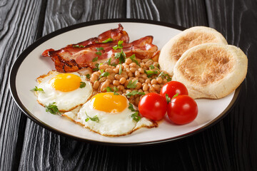 Delicious breakfast of fried eggs, beans, bacon, tomatoes and English muffins close-up in a plate on the table. horizontal