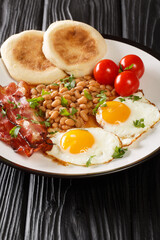 Hot hearty breakfast of fried eggs, beans, bacon, tomatoes and muffins close-up in a plate on the table. vertical