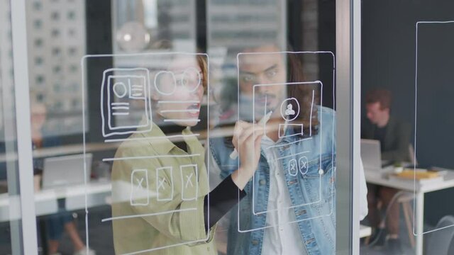 PAN shot of young female UI designer drawing interface ideas on glass wall or window in office and talking to male colleague