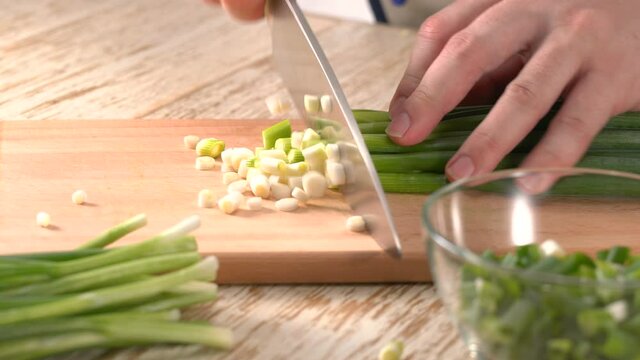 Close up shot of chef's hands using a knife cutting a fresh green onion or spring onion on wooden board. 