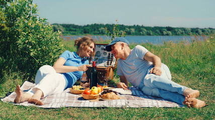 A happy, slender elderly couple, a man and a woman, are lying on the grass in a park by the river and are having an exciting conversation, smiling on a sunny summer day.