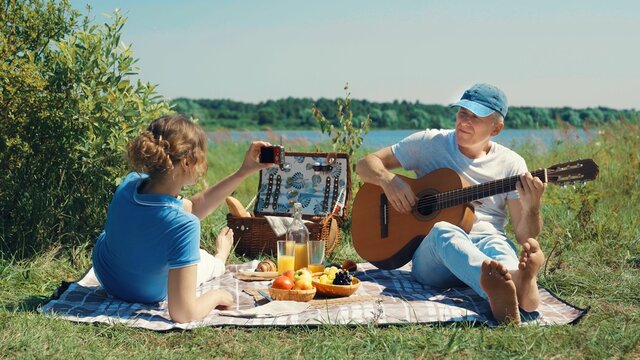 A tourist elderly slender couple arranges a romantic picnic on the beach on a sunny summer day: a man smiles and plays the guitar for a friend, and she takes pictures of him with a camera.