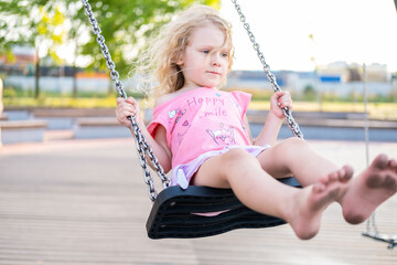 little cute caucasian curly-haired girl swinging on a swing in the park with a sad face and dirty feet. concept childhood.