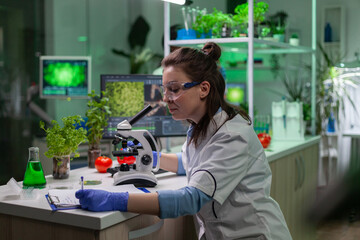 Biologist scientist looking at leaf sample using medical microscope while writing gmo expertise on notepad. Chemist examining biological discovery on plant working in pharmaceutical laboratory.