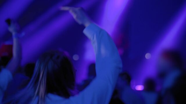 Slow motion: long hair teenage girl partying, cheering, raising hands up and jumping at rock concert in front of stage of nightclub. Bright colorful stage lighting. Nightlife and entertainment concept