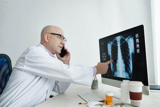 Mature doctor sitting at the table and pointing at computer monitor with x-ray image and discussing it on mobile phone
