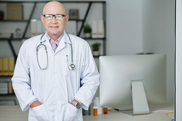 Portrait of mature professional doctor in white coat looking at camera while standing at his office