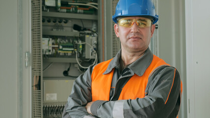 Portrait of mature employee in helmet and protective goggles standing with crossed arms near switchboard at production plant close view