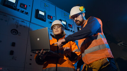 Engineers bearded man and woman in orange uniform and helmets use laptop to check equipment in control room of production plant low angle shot